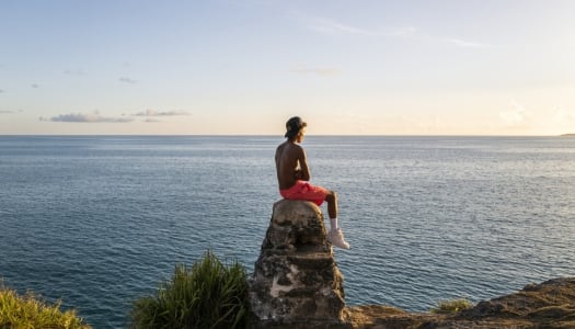 A young boy is sitting on a rock looking out at a calm ocean.